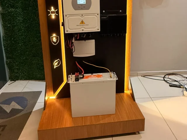 Kit de demonstração com inversor híbrido e bateria de armazenamento exibido em estande da Eletroserv, com tela mostrando instalação solar