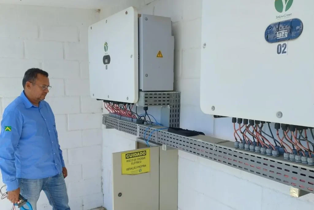 Técnico observando painel de inversores solares instalados em parede, com sinalização de segurança de geração própria