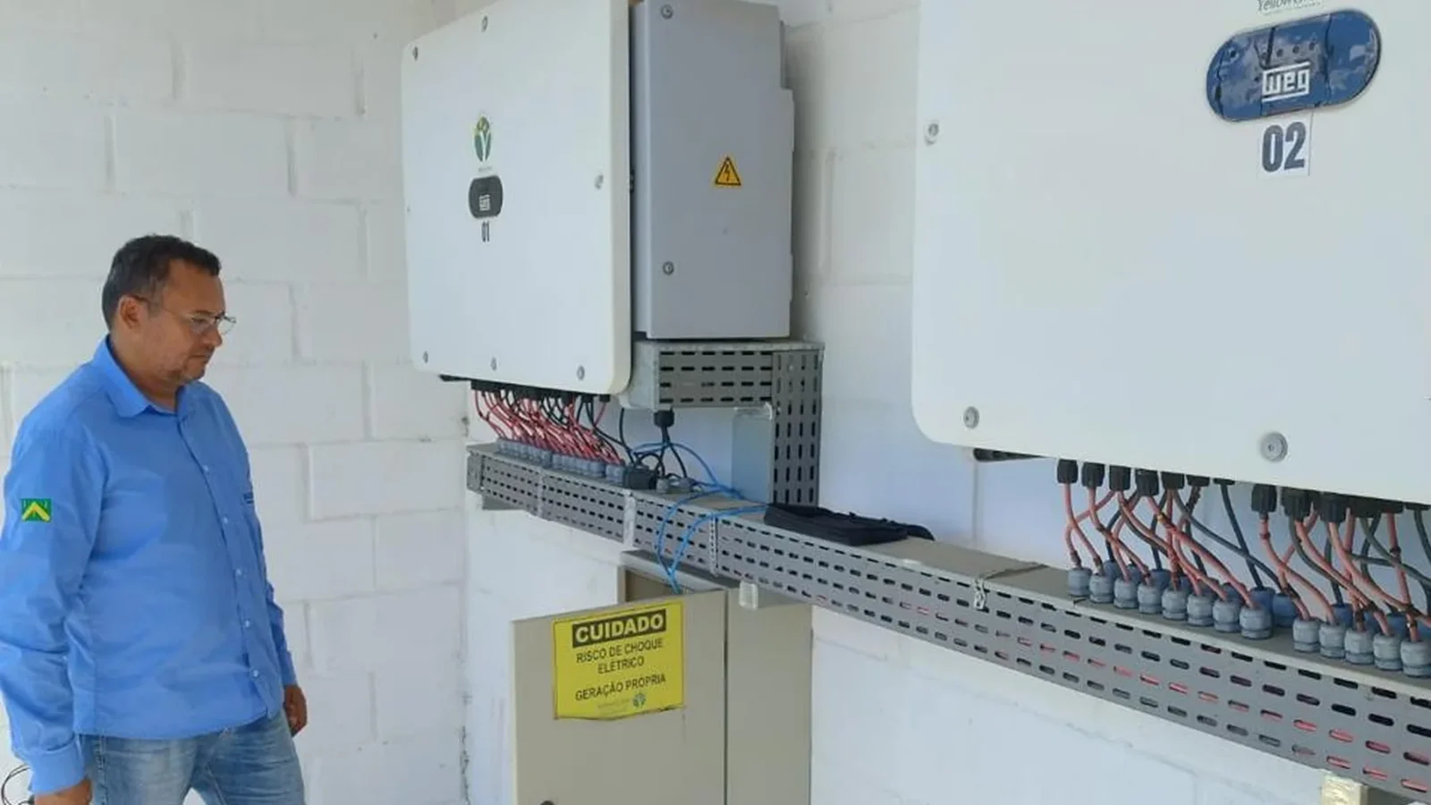 Técnico observando painel de inversores solares instalados em parede, com sinalização de segurança de geração própria