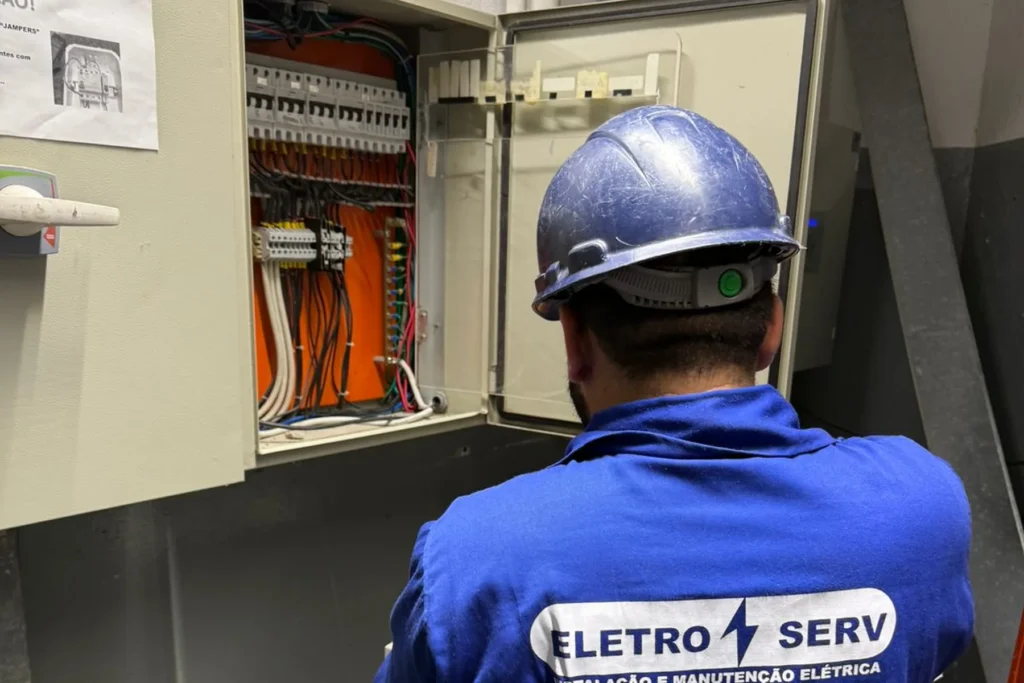 Técnico uniformizado com capacete inspeciona quadro de distribuição elétrica aberto em ambiente industrial.