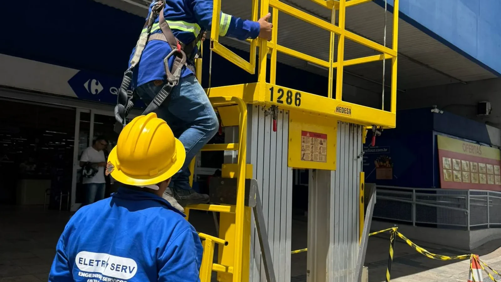 Dois técnicos uniformizados sobem em plataforma elevatória amarela em frente a loja Carrefour, durante o dia.