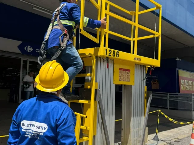 Dois técnicos uniformizados sobem em plataforma elevatória amarela em frente a loja Carrefour, durante o dia.