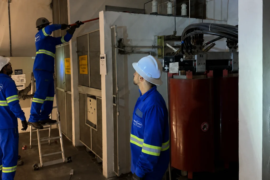 Três técnicos realizam serviço de manutenção em sala de subestação, um deles em escada próximo a painel elétrico, ao lado de transformador a seco.