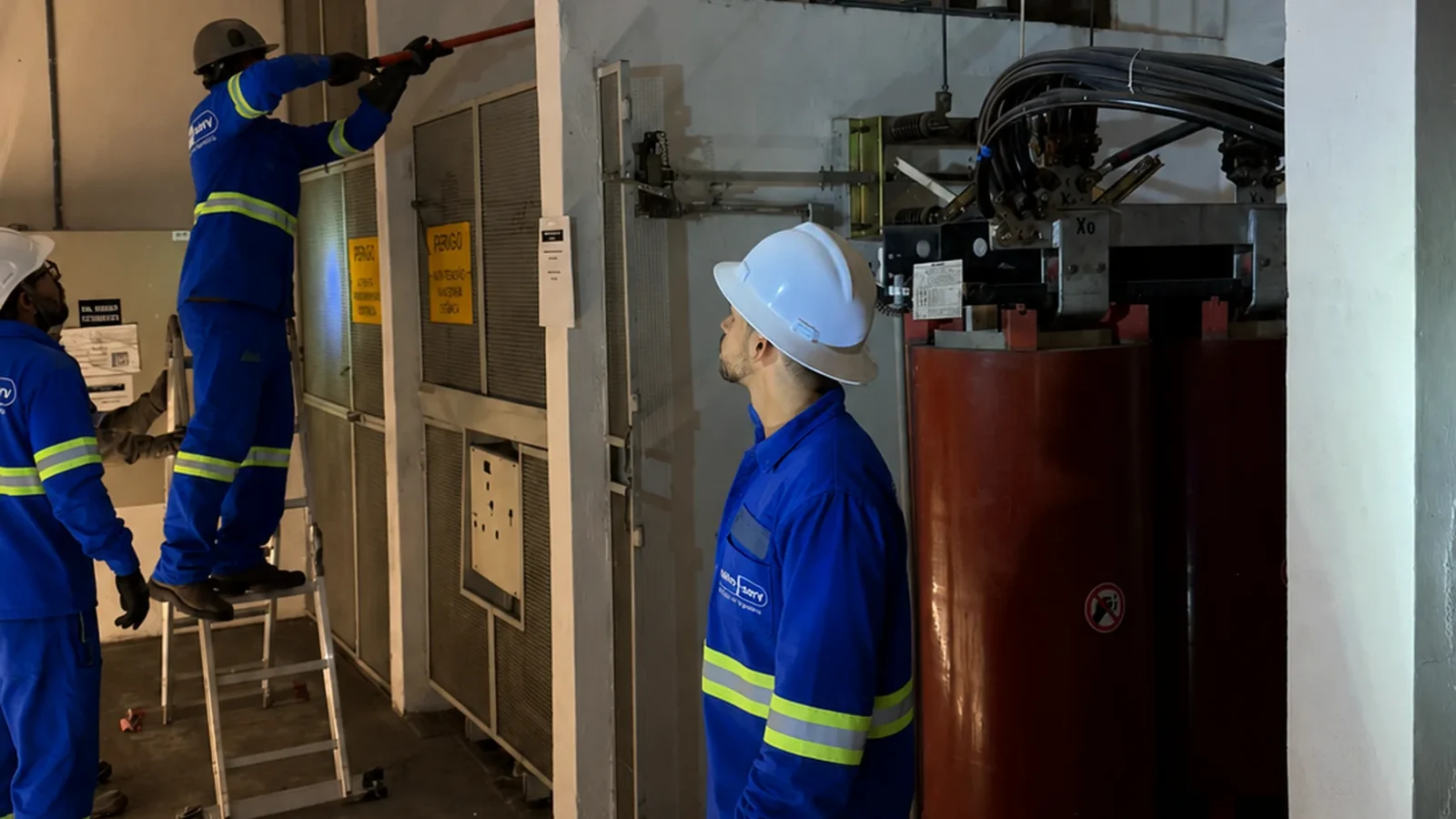 Três técnicos realizam serviço de manutenção em sala de subestação, um deles em escada próximo a painel elétrico, ao lado de transformador a seco.