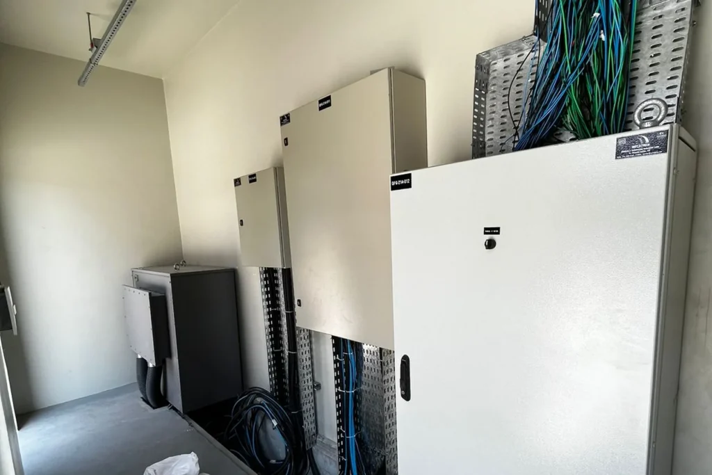 Corredor de sala técnica com três quadros elétricos instalados na parede e cabos organizados em leito aéreo.