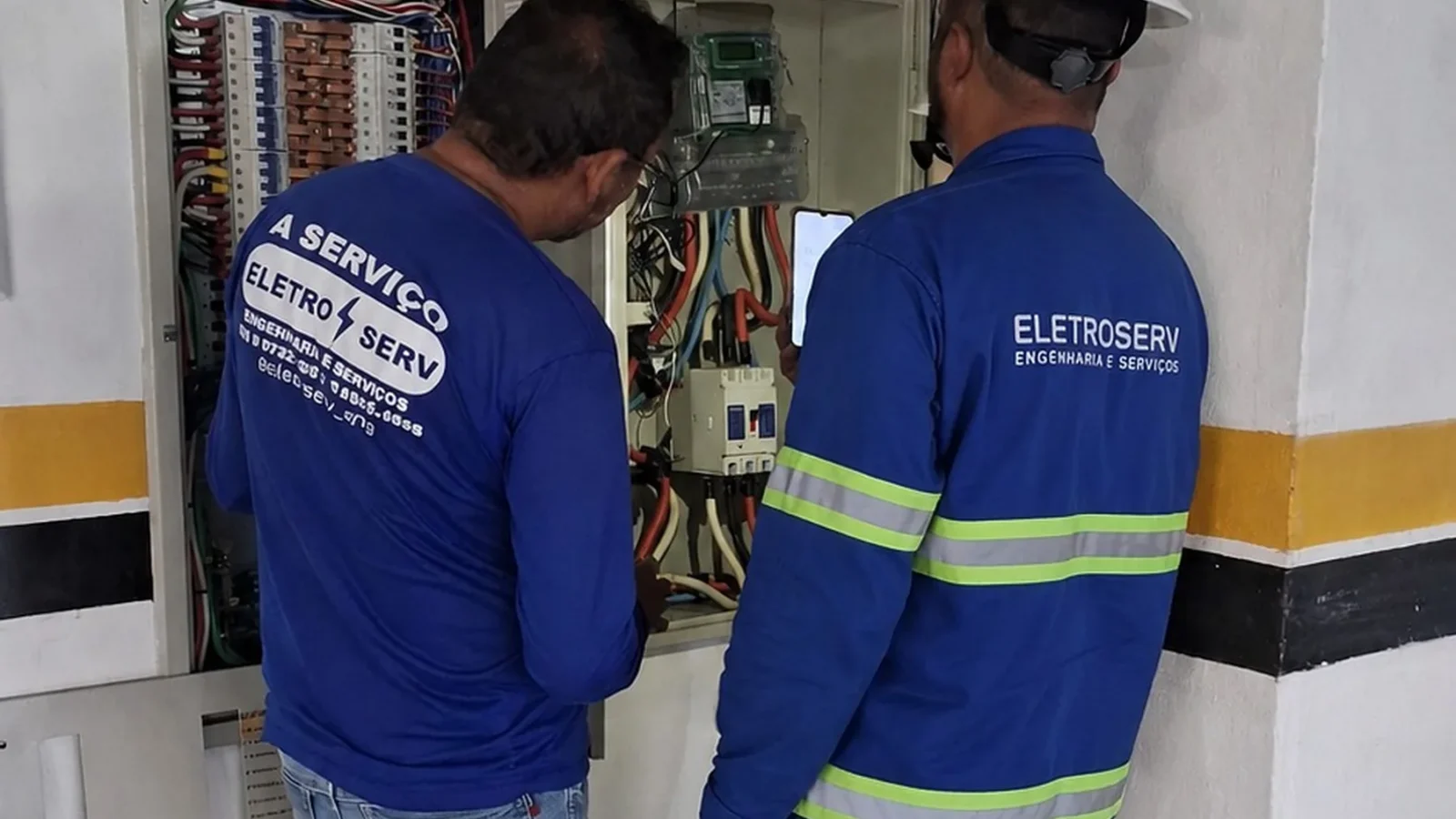 Dois técnicos uniformizados observam quadro elétrico aberto, um deles consultando informações no celular.