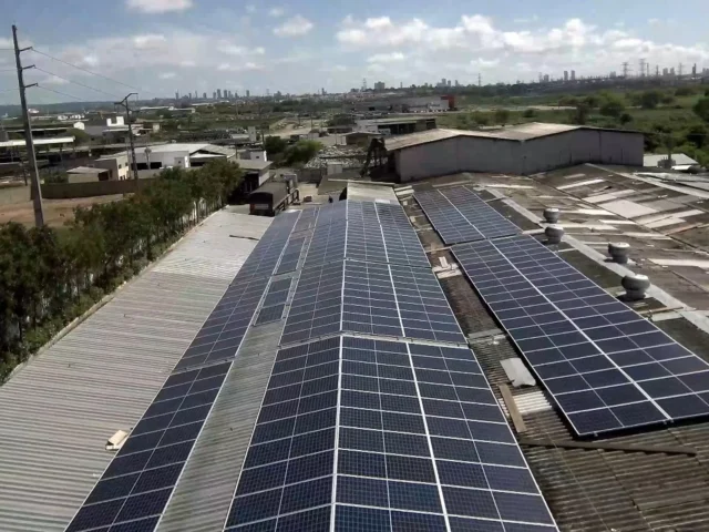 Vista aérea de painéis solares instalados sobre telhados de galpões industriais, com área urbana ao fundo.
