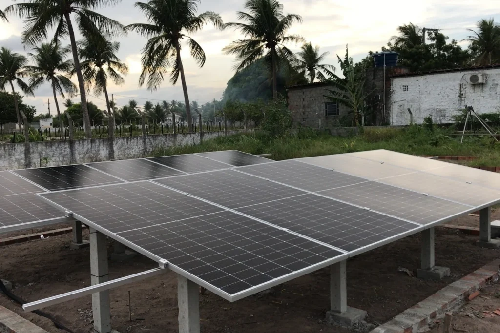 Painéis solares montados em estrutura elevada em quintal cercado por coqueiros, próximo a casa de alvenaria, ao entardecer.