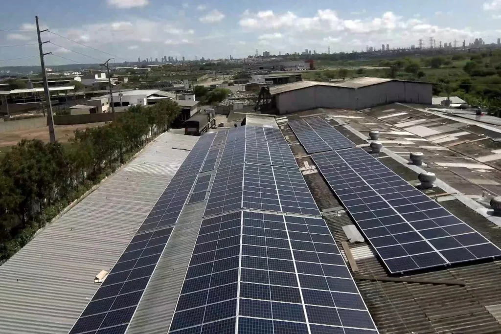 Vista aérea de painéis solares instalados sobre telhados de galpões industriais, com área urbana ao fundo.