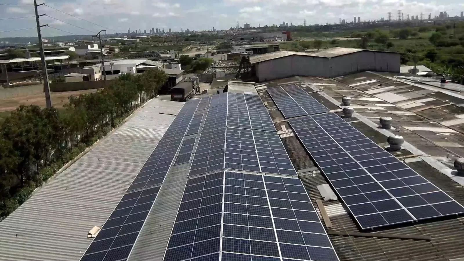 Vista aérea de painéis solares instalados sobre telhados de galpões industriais, com área urbana ao fundo.