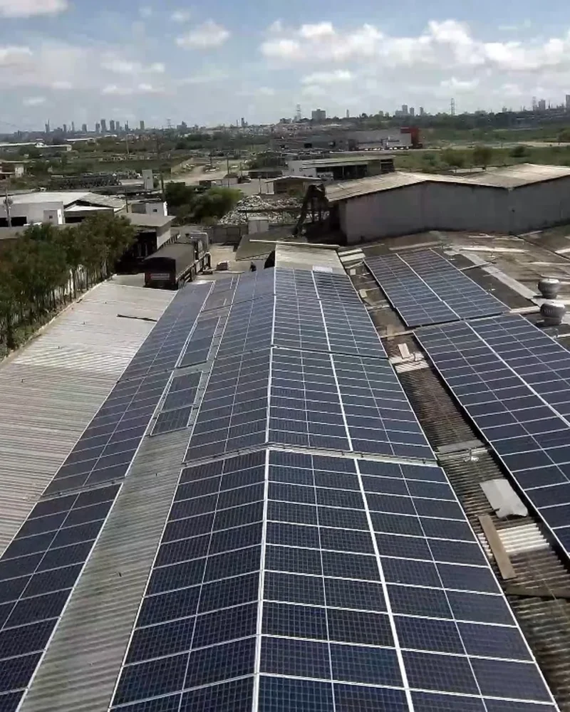 Vista aérea de painéis solares instalados sobre telhados de galpões industriais, com área urbana ao fundo.