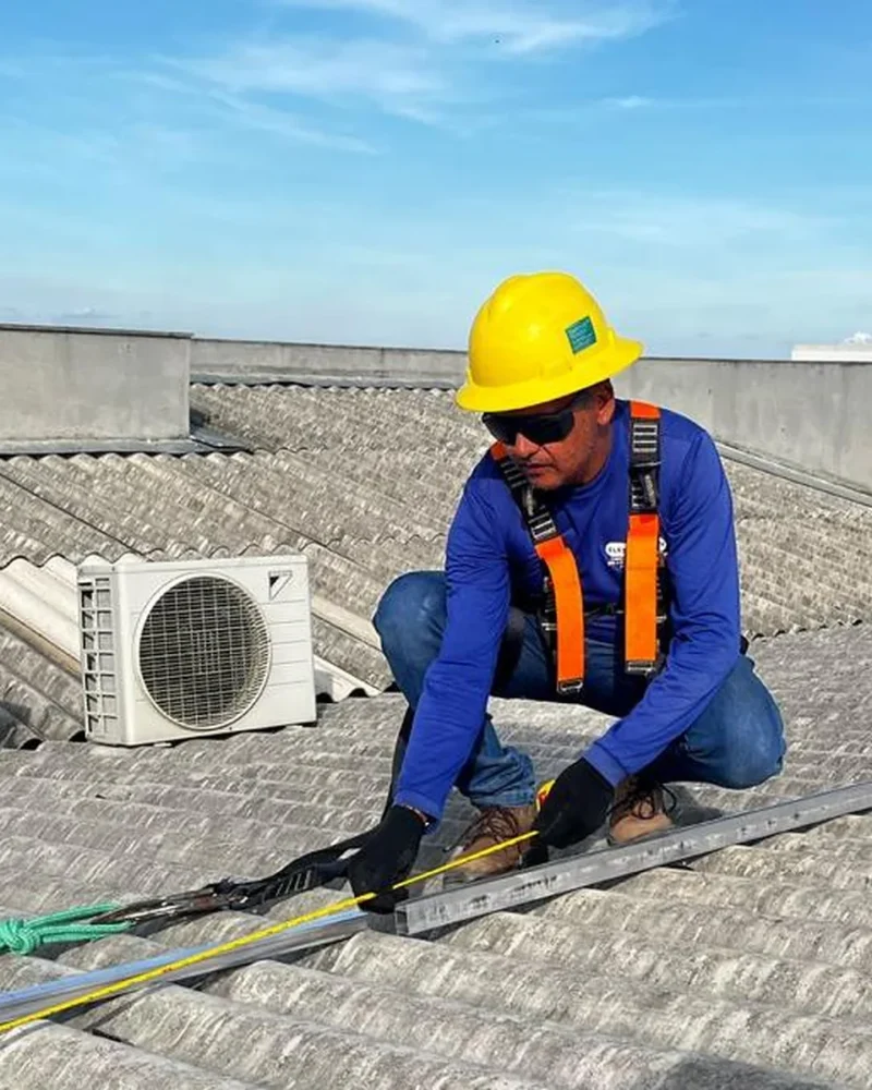 Técnico agachado fixa trilho metálico de linha de vida em telhado de fibrocimento, usando equipamento de proteção contra quedas.