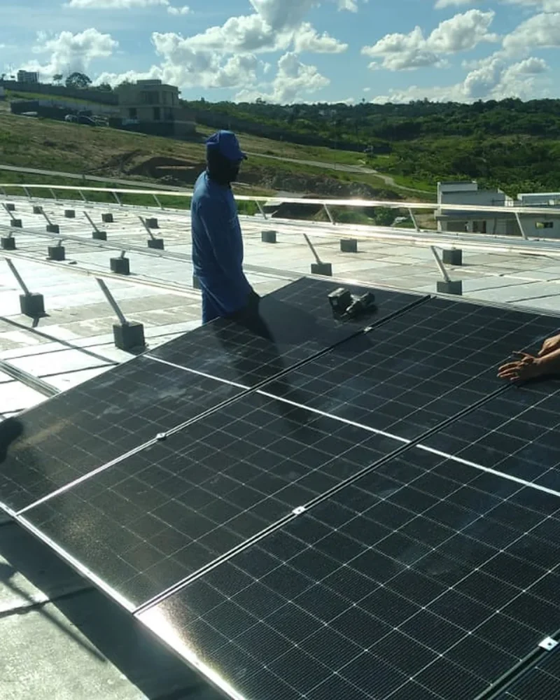 Três técnicos posicionam painel solar em estrutura de telhado, com colinas verdes ao fundo em dia ensolarado.