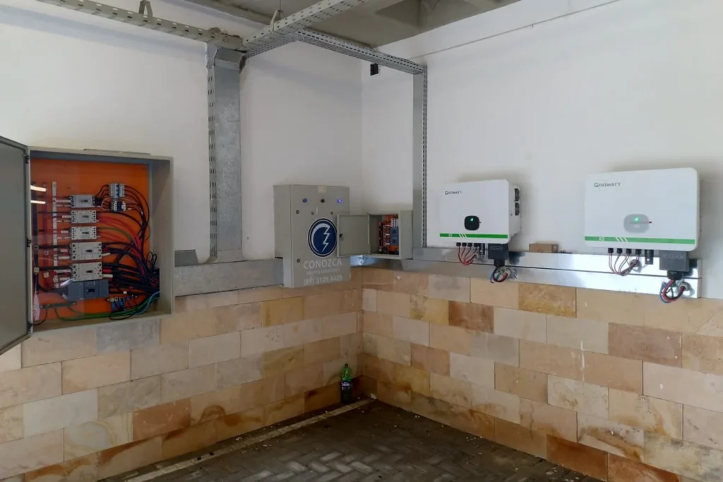 Ambiente técnico com quadro elétrico aberto, painel de controle e dois inversores solares montados na parede.