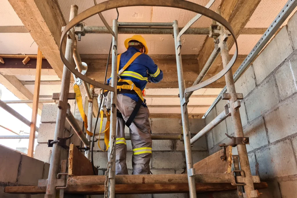 Técnico sobre andaime realiza instalação elétrica junto à laje, dentro de ambiente em construção com paredes de bloco aparente.