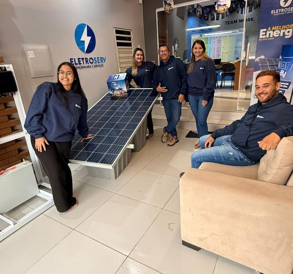 Equipe da Eletroserv reunida em escritório, ao lado de painel solar exposto e banners promocionais da empresa