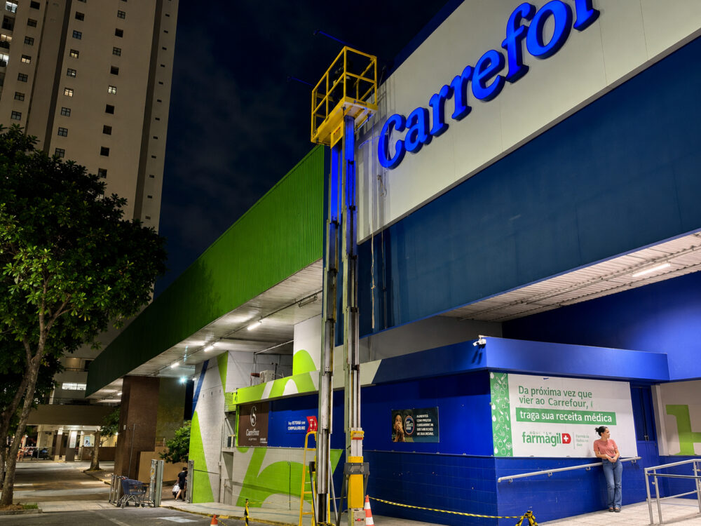 Técnico da Eletroserv observa equipe posicionando poste de concreto com auxílio de guindaste em canteiro de obras, com prédios ao fundo.