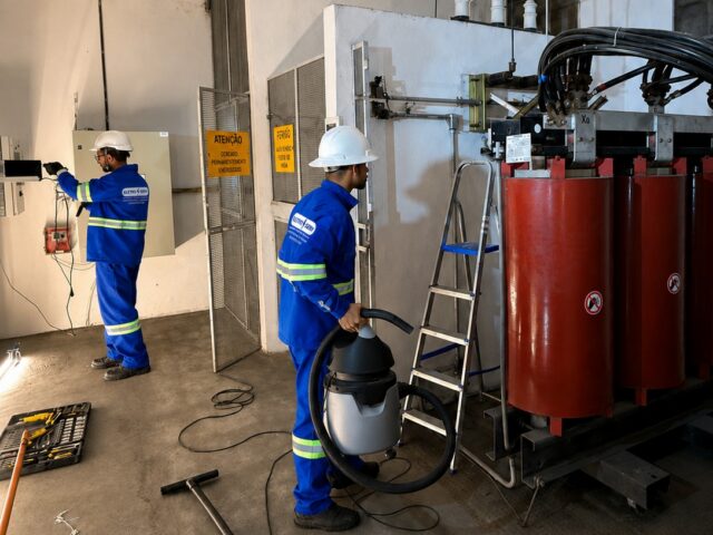 Dois técnicos uniformizados realizam limpeza em transformador a seco dentro de sala de subestação, um deles usando aspirador industrial.